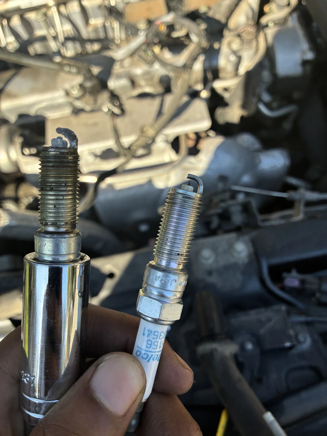 Old and new spark plugs held side by side in front of an engine